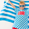 Child wearing a blue and white checkered shirt with 'USA' printed on it, standing against a blue and white striped towel fabric.
