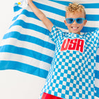 Child wearing a blue and white checkered shirt with 'USA' printed on it, standing against a blue and white striped towel fabric.