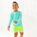 Child wearing neon yellow swim shorts with blue lightning bolt pattern and turquoise rash guard on a white background