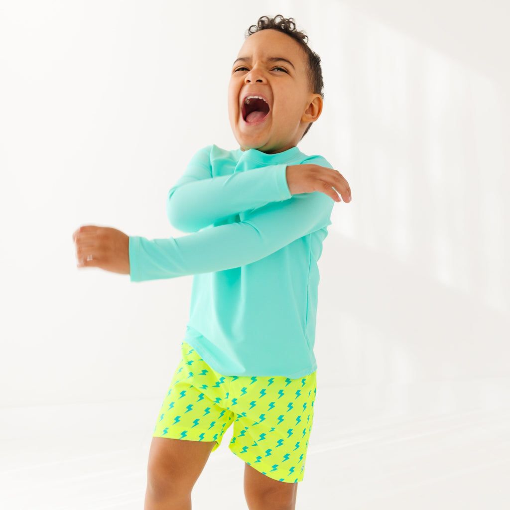 Child wearing neon yellow swim shorts with blue lightning bolt pattern and turquoise rash guard on a white background
