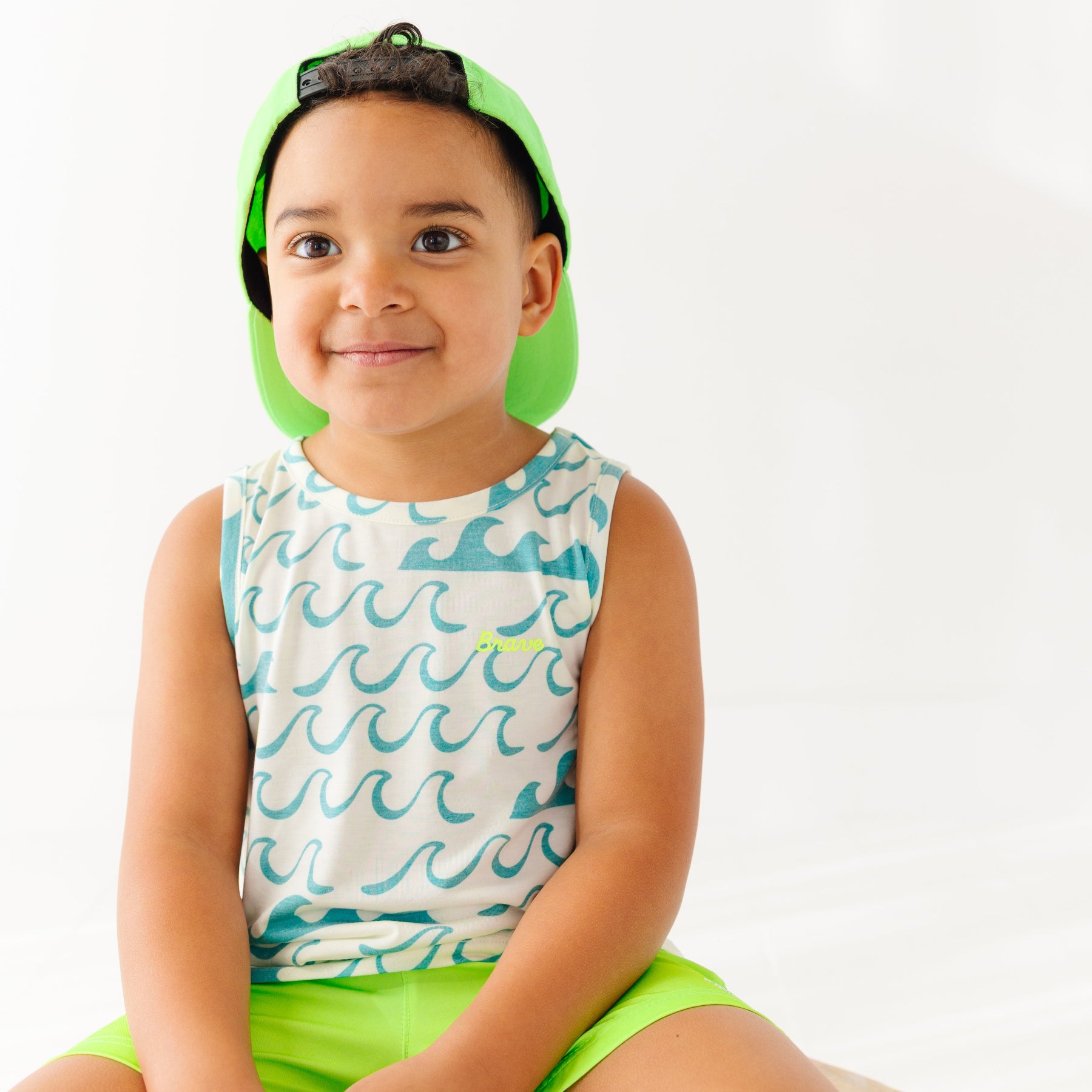 Child wearing a tank top with waves pattern, green swim trunks and green trucker hat on a white background