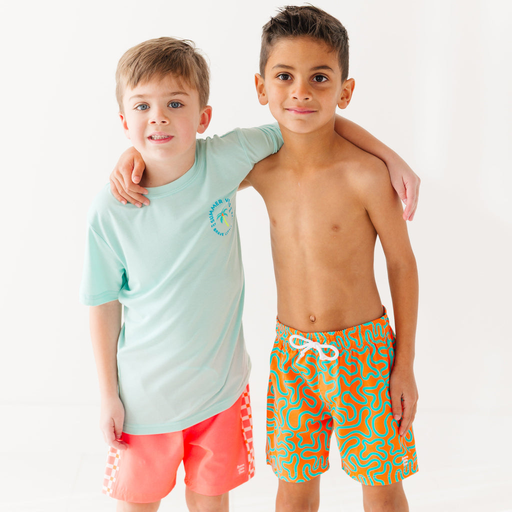 Child wearing orange and blue squiggly-patterned swim shorts and another child wearing a blue summer shirt and coral checkered swim shorts