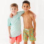 Child wearing orange and blue squiggly-patterned swim shorts and another child wearing a blue summer shirt and coral checkered swim shorts
