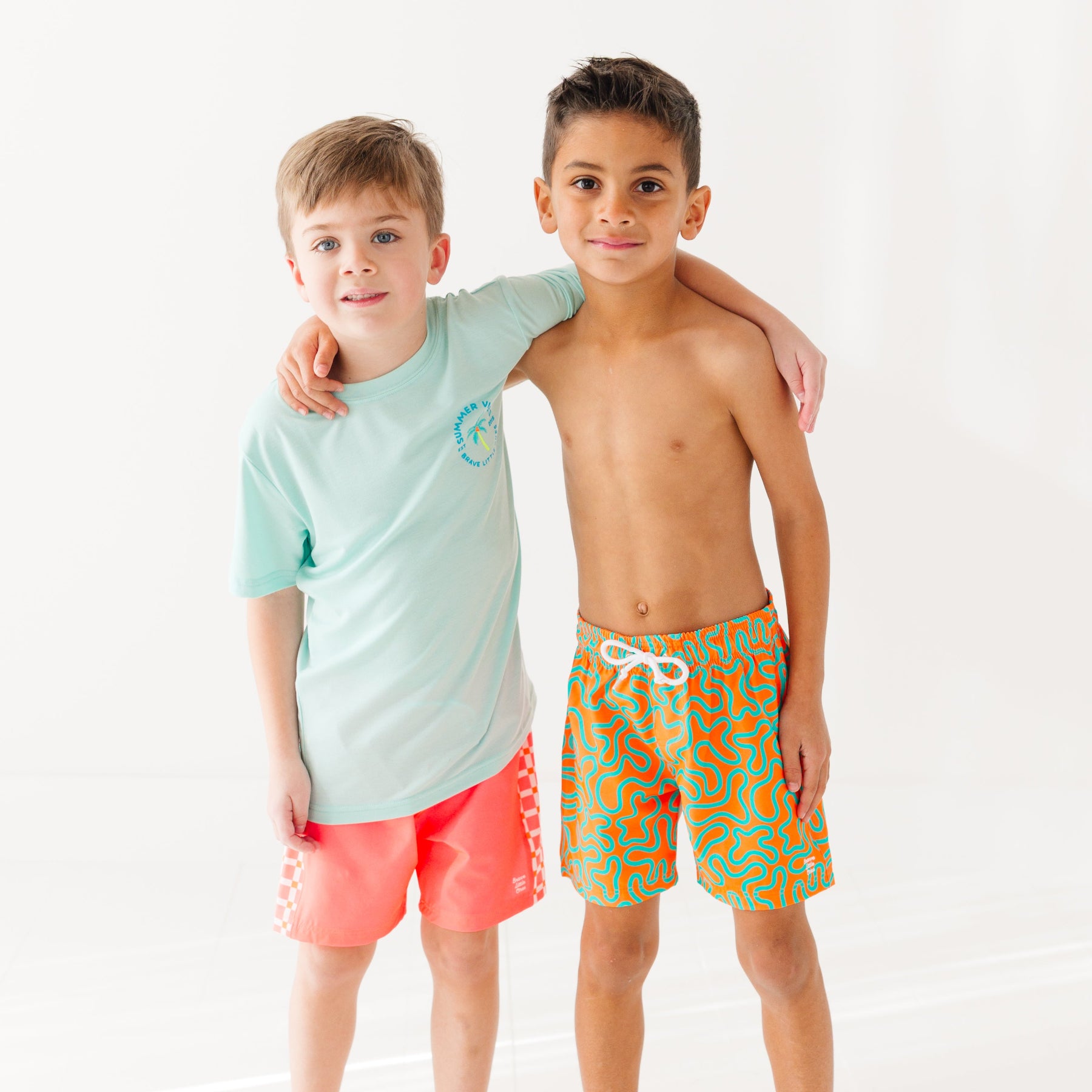 Child wearing orange and blue squiggly-patterned swim shorts and another child wearing a blue summer shirt and coral checkered swim shorts