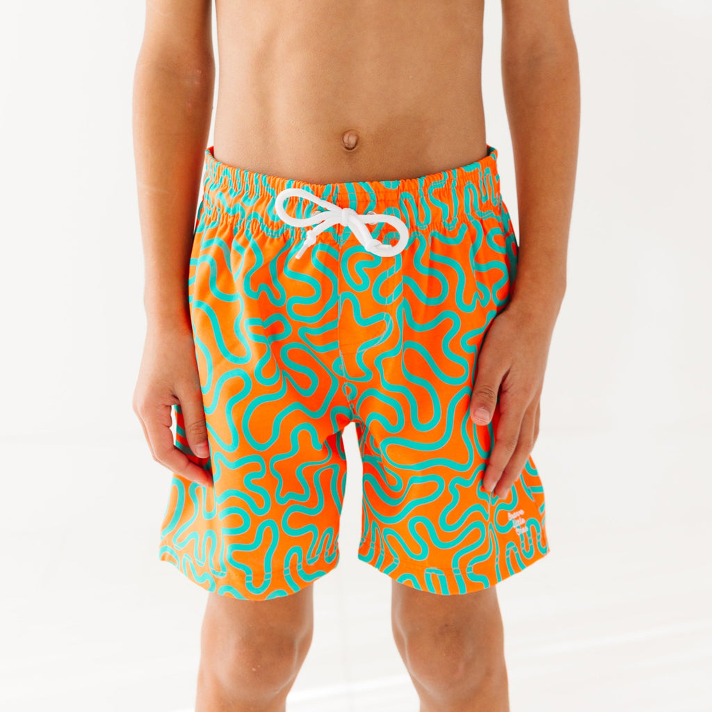 Close up of a child wearing orange and blue squiggly-patterned swim shorts on a white background