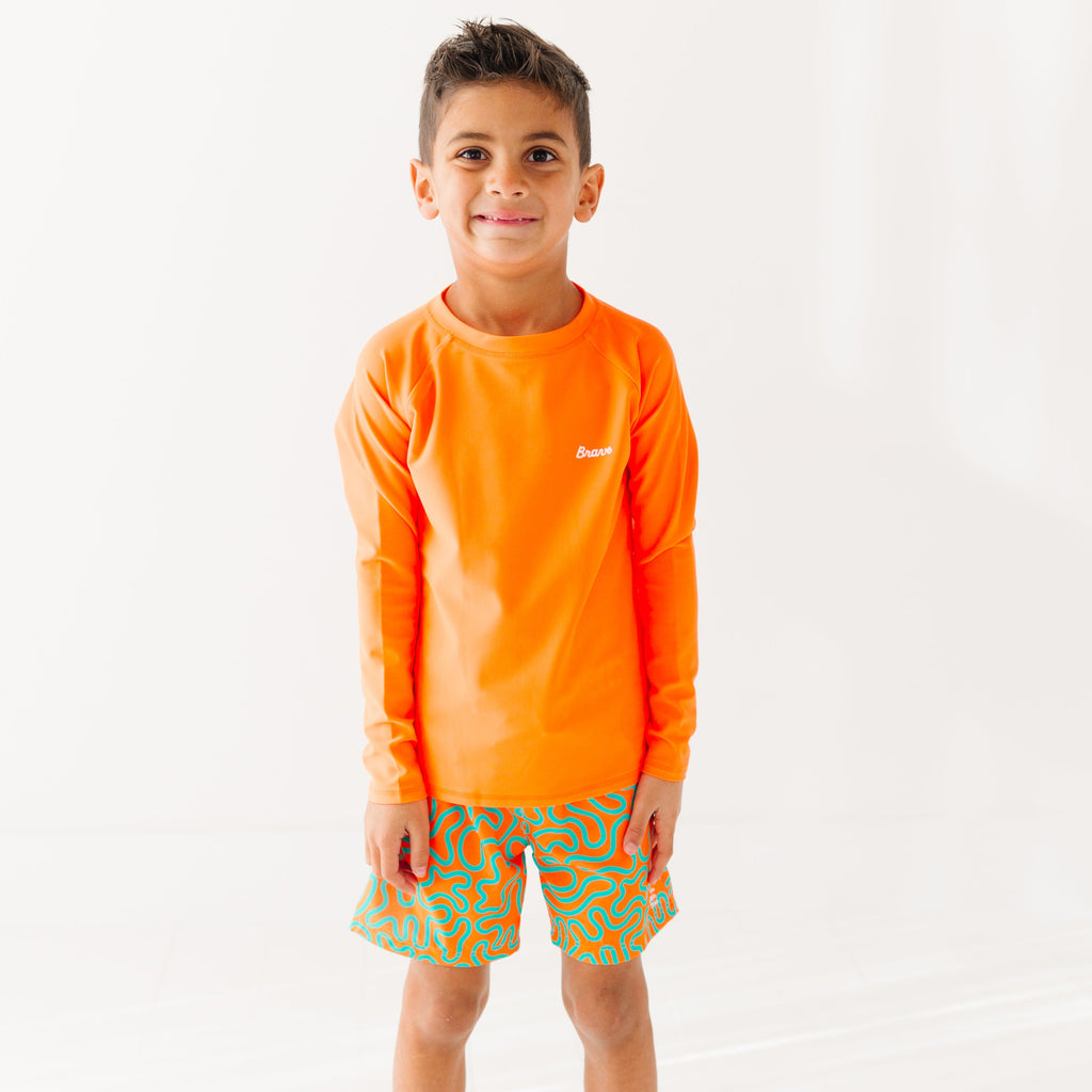 Child wearing orange and blue squiggly-patterned swim shorts and matching orange rash guard on a white background