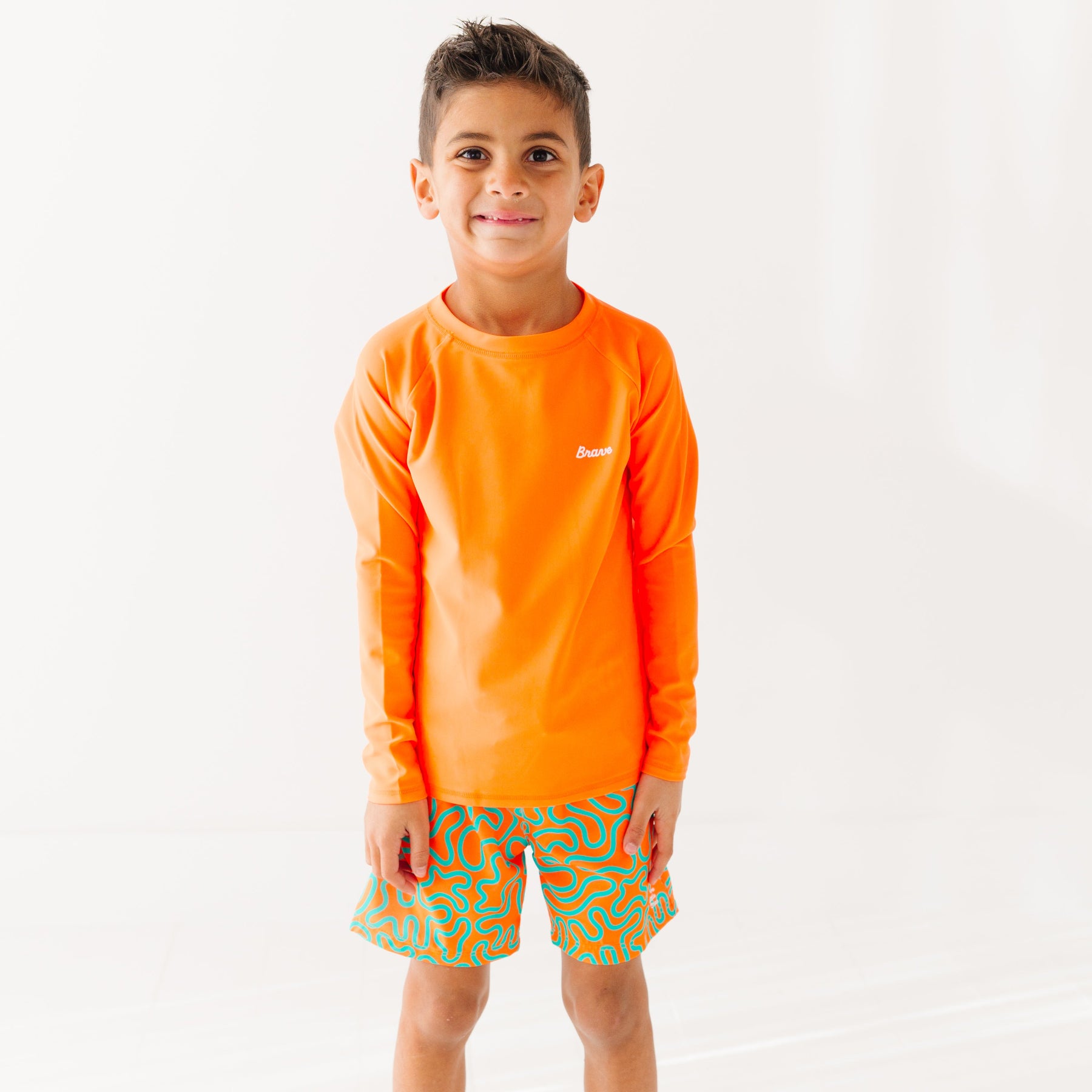 Child wearing orange and blue squiggly-patterned swim shorts and matching orange rash guard on a white background