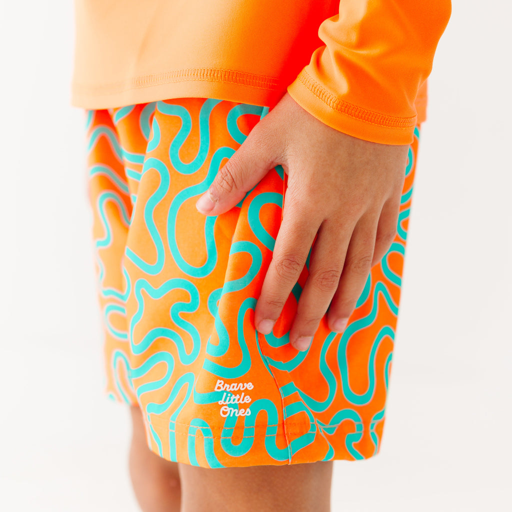 Close up shot of a child wearing orange and blue squiggly-patterned swim shorts and matching orange rash guard on a white background