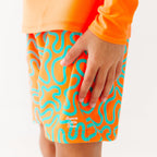 Close up shot of a child wearing orange and blue squiggly-patterned swim shorts and matching orange rash guard on a white background
