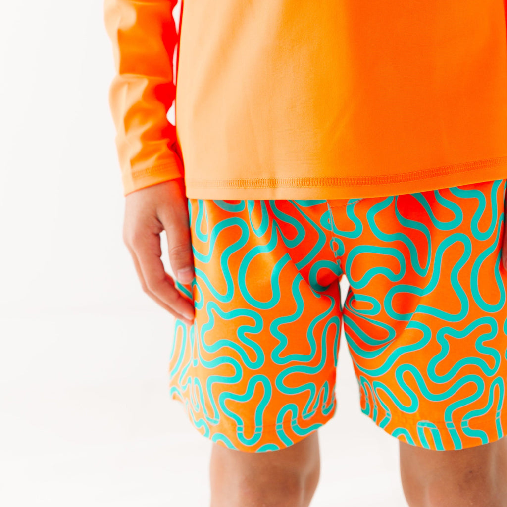 Close up of a child wearing orange and blue squiggly-patterned swim shorts and matching orange rash guard on a white background