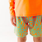 Close up of a child wearing orange and blue squiggly-patterned swim shorts and matching orange rash guard on a white background