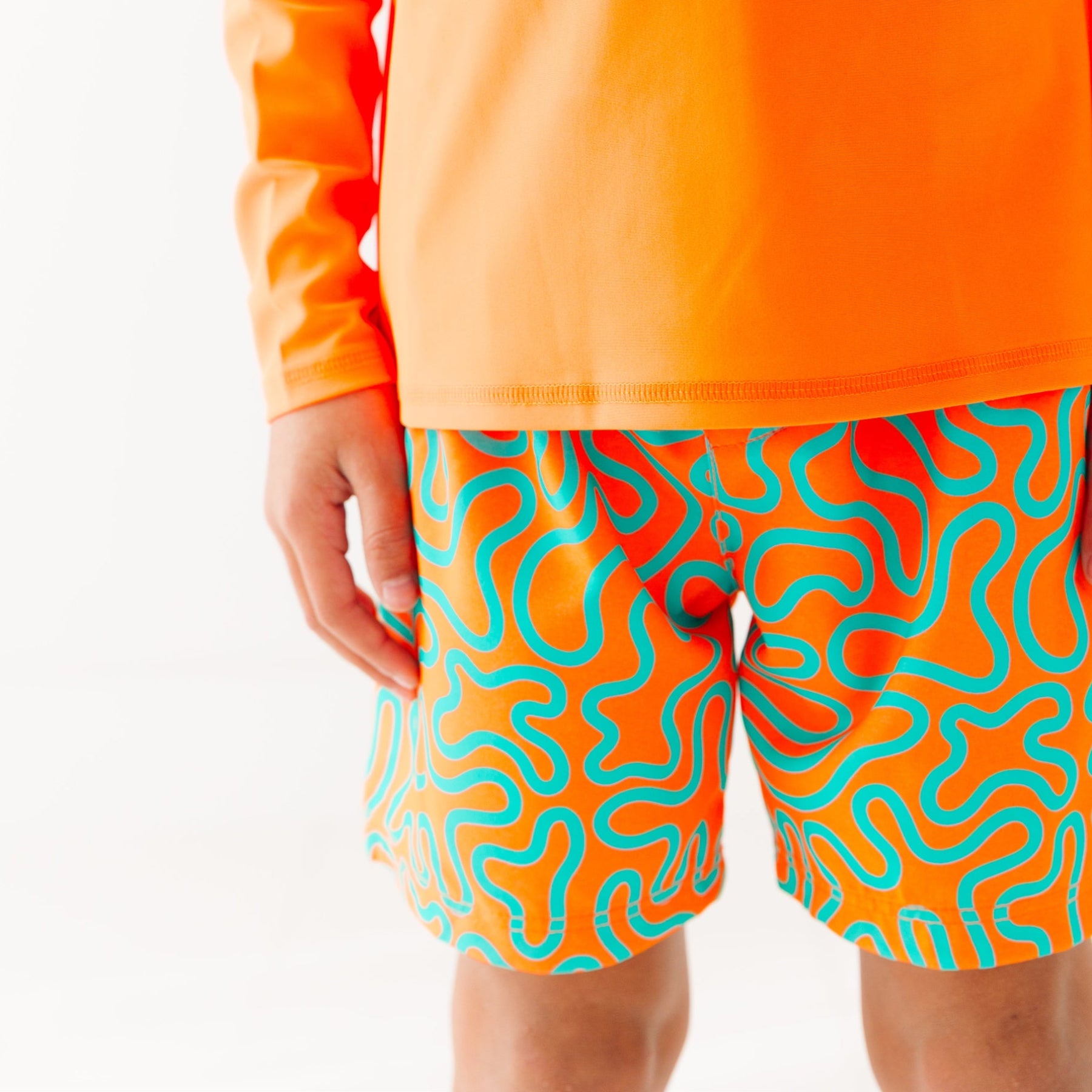 Close up of a child wearing orange and blue squiggly-patterned swim shorts and matching orange rash guard on a white background