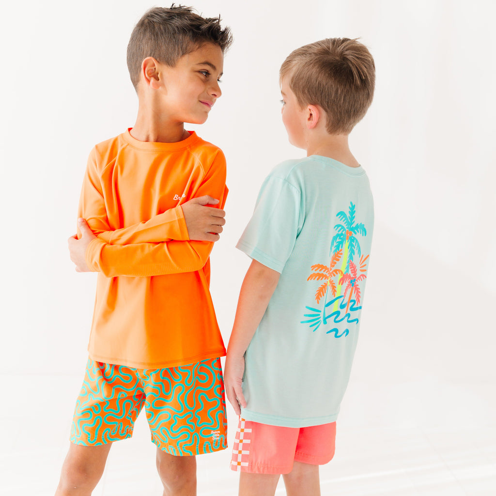 Child wearing orange and blue squiggly-patterned swim shorts and orange rash guard, and another child wearing a blue summer shirt and coral checkered swim shorts