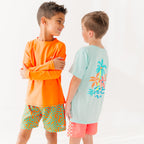 Child wearing orange and blue squiggly-patterned swim shorts and orange rash guard, and another child wearing a blue summer shirt and coral checkered swim shorts