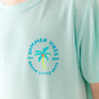 Close up shot of a light blue t-shirt with 'Summer Vibes' text and palm tree design on a plain background