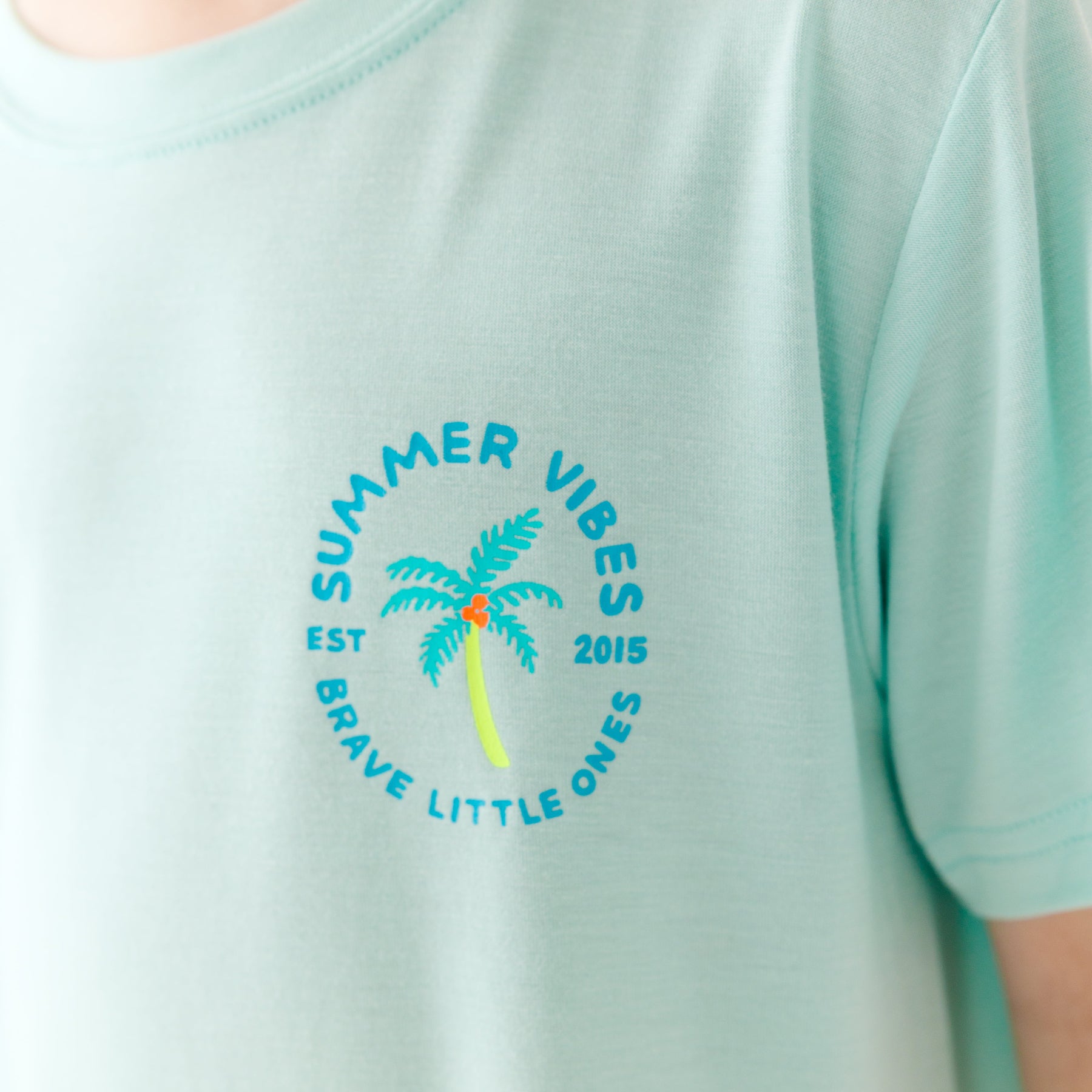 Close up shot of a light blue t-shirt with 'Summer Vibes' text and palm tree design on a plain background