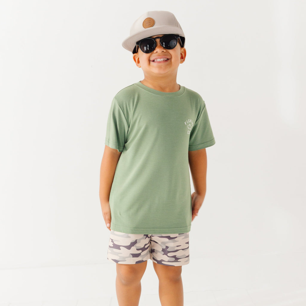 Child wearing a green t-shirt with a graphic, camouflage shorts, sunglasses and taupe cap on a white background