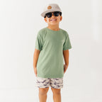 Child wearing a green t-shirt with a graphic, camouflage shorts, sunglasses and taupe cap on a white background
