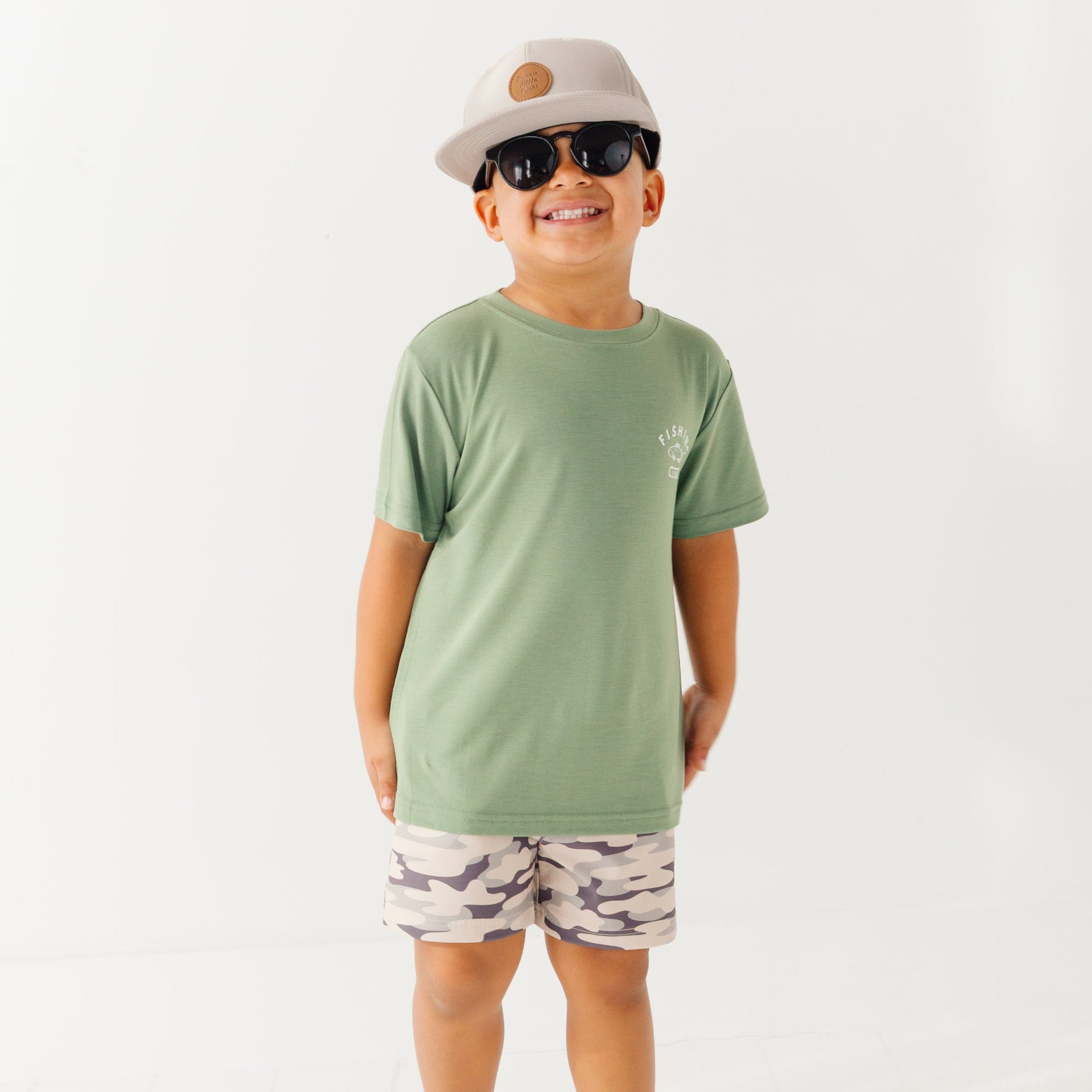 Child wearing a green t-shirt with a graphic, camouflage shorts, sunglasses and taupe cap on a white background