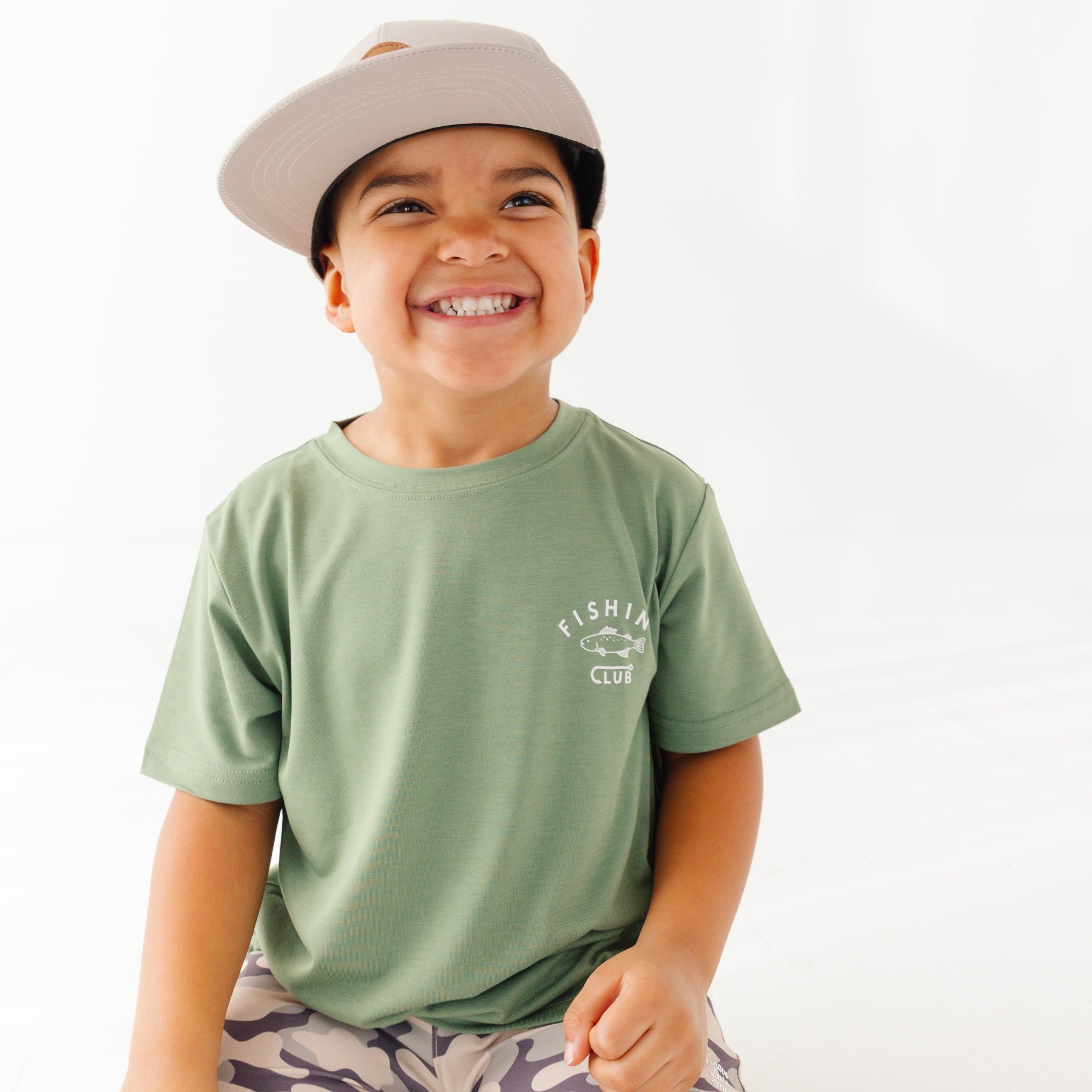 Child wearing a green t-shirt with a graphic, camouflage shorts and taupe cap on a white background