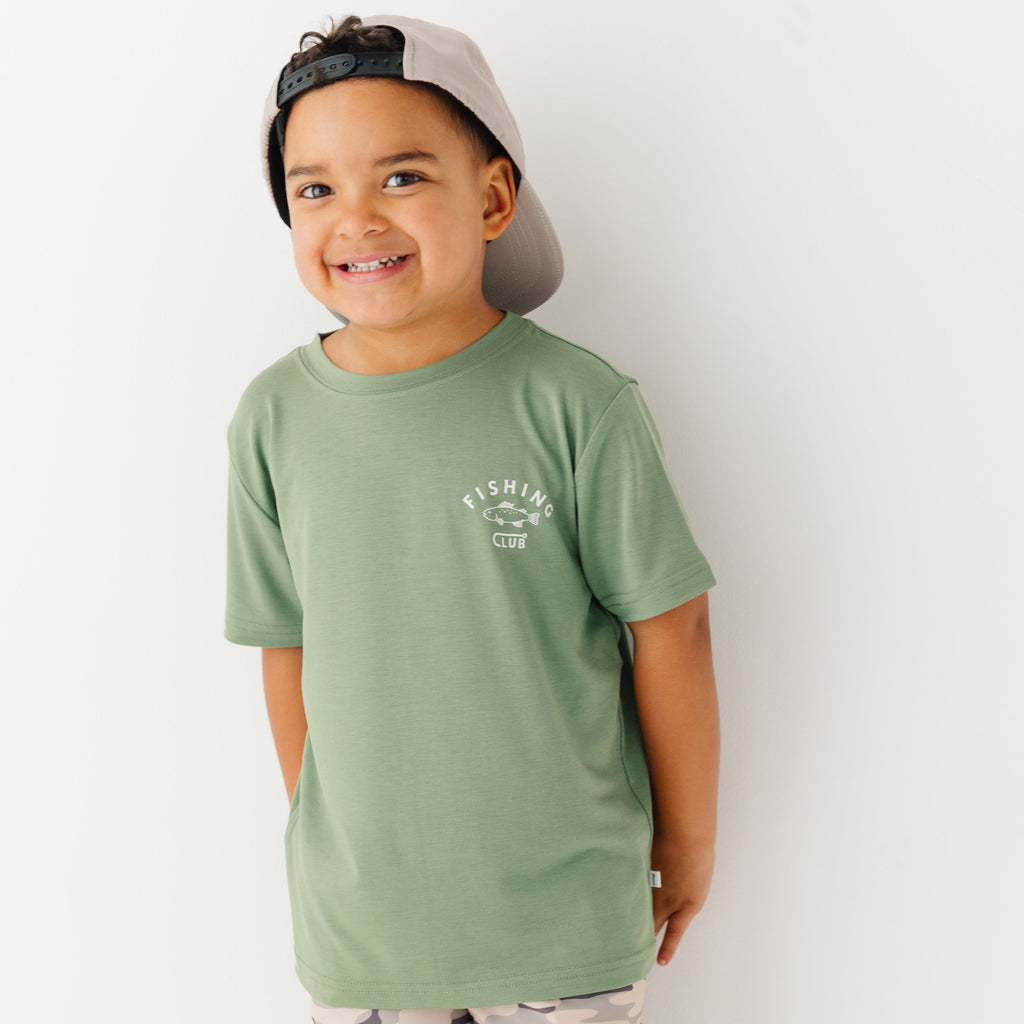 Child wearing a green t-shirt with a graphic and taupe cap on a white background