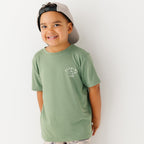 Child wearing a green t-shirt with a graphic and taupe cap on a white background