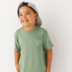 Child wearing a green t-shirt with a graphic and taupe cap on a white background
