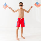 Child wearing red swim shorts with a flag design, holding two American flags on a white background