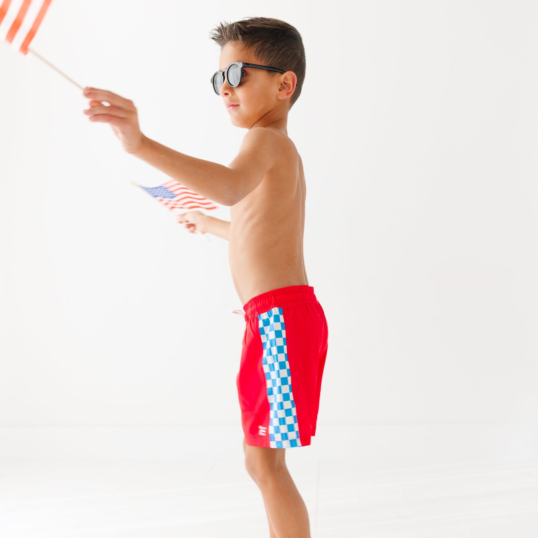 Young boy holding American flags, wearing red swim shorts with a checkered pattern, on a white background.