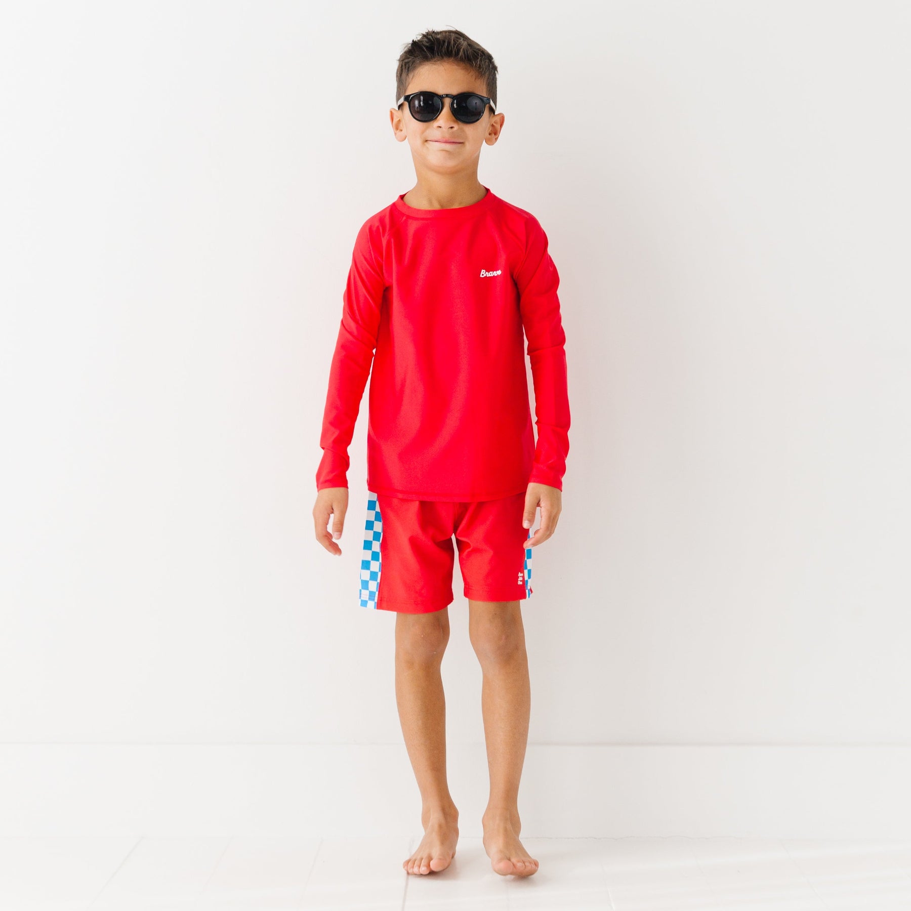 Child wearing a red rash guard, matching red swim trunks and sunglasses on a white background