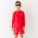 Child wearing a red rash guard, matching red swim trunks and sunglasses on a white background