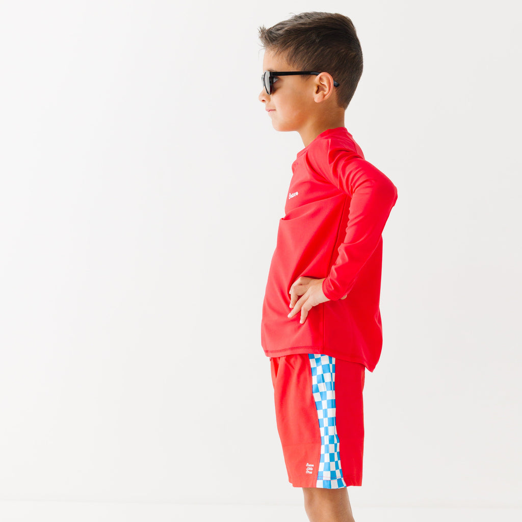 Child wearing a red rash guard, matching red swim trunks and sunglasses on a white background