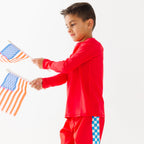 Child wearing a red rash guard, matching red swim trunks, holding small American flags on a white background