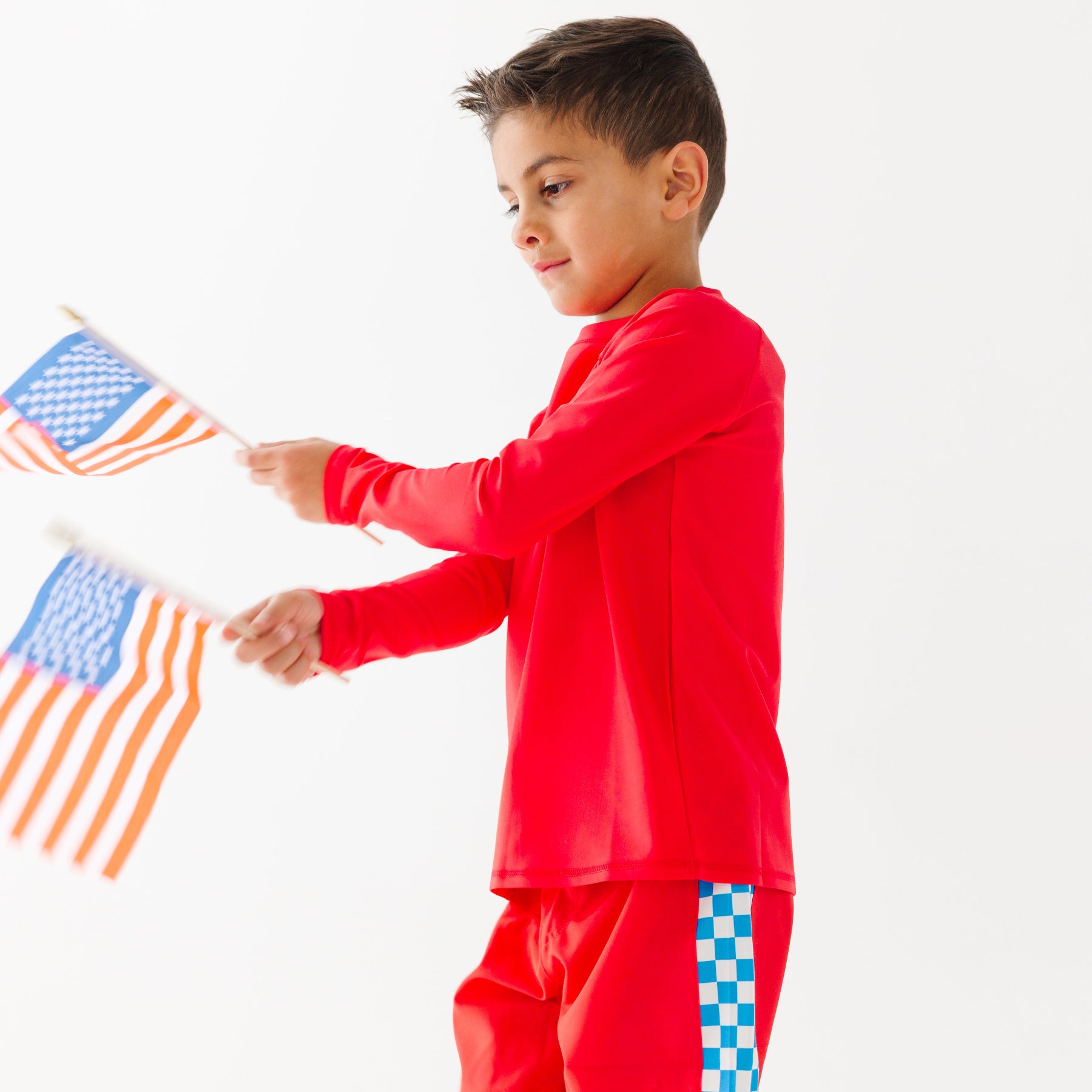 Child wearing a red rash guard, matching red swim trunks, holding small American flags on a white background