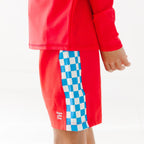Close up shot of a child wearing a red long-sleeve rash guard and shorts with checkered pattern on a white background