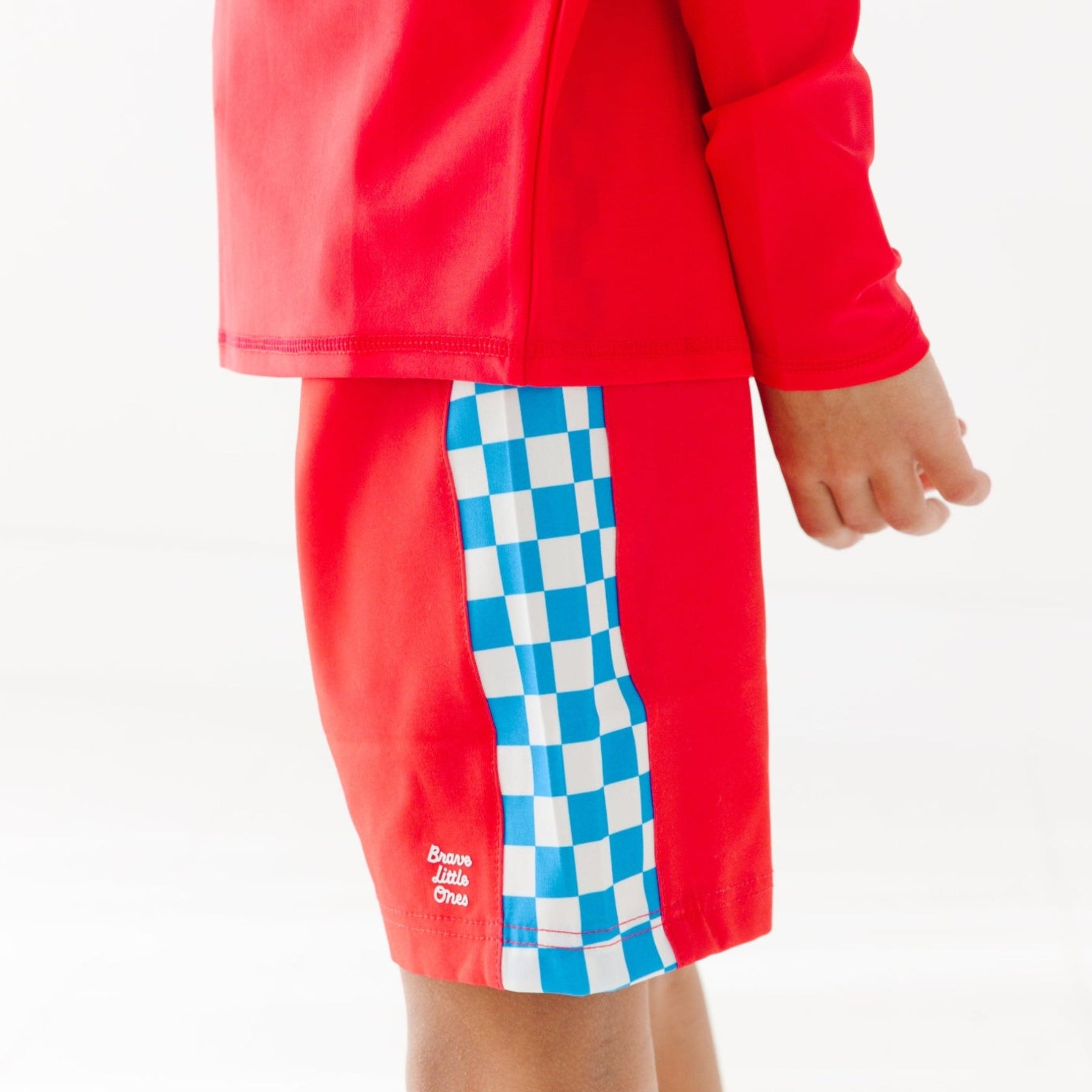 Close up shot of a child wearing a red long-sleeve rash guard and shorts with checkered pattern on a white background