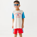 Child wearing a white t-shirt with blue sleeves and red shorts, wearing sunglasses on a white background