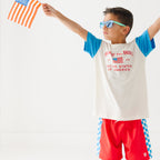 Child wearing a white t-shirt with blue sleeves and red shorts, holding a small American flag on a white background