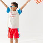 Child wearing a white t-shirt with blue sleeves and red shorts, holding a small American flag on a white background