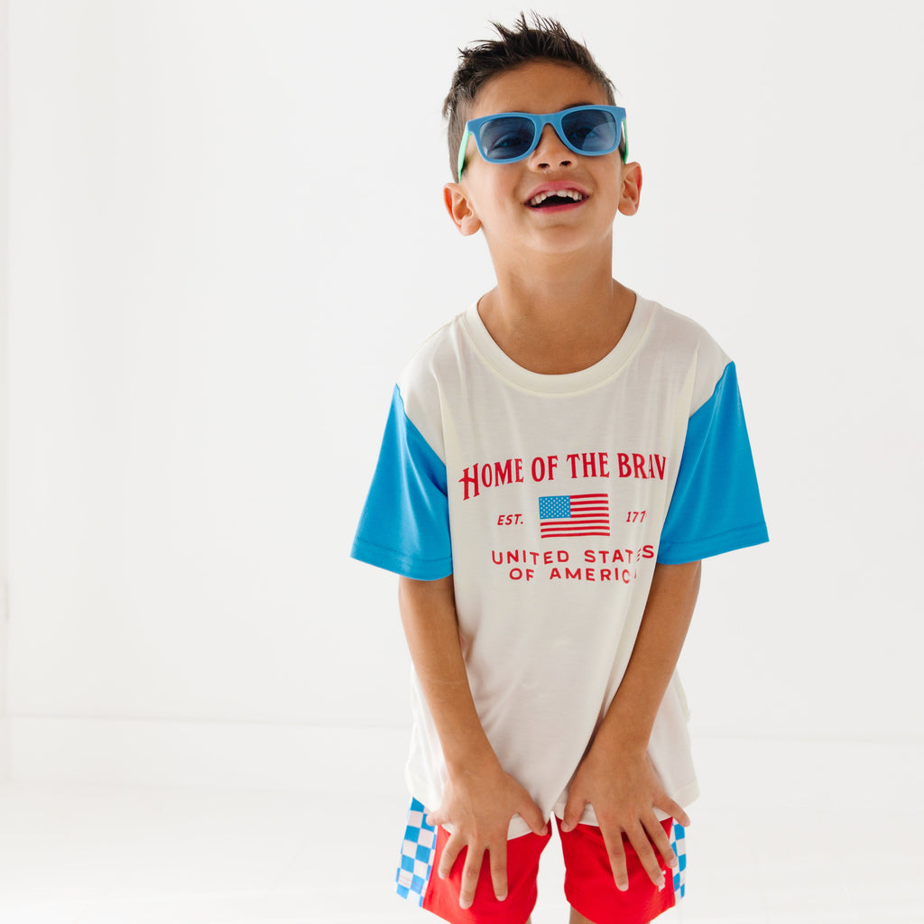 Child wearing a white t-shirt with blue sleeves, red shorts and blue sunglasses on a white background