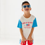 Child wearing a white t-shirt with blue sleeves, red shorts and blue sunglasses on a white background