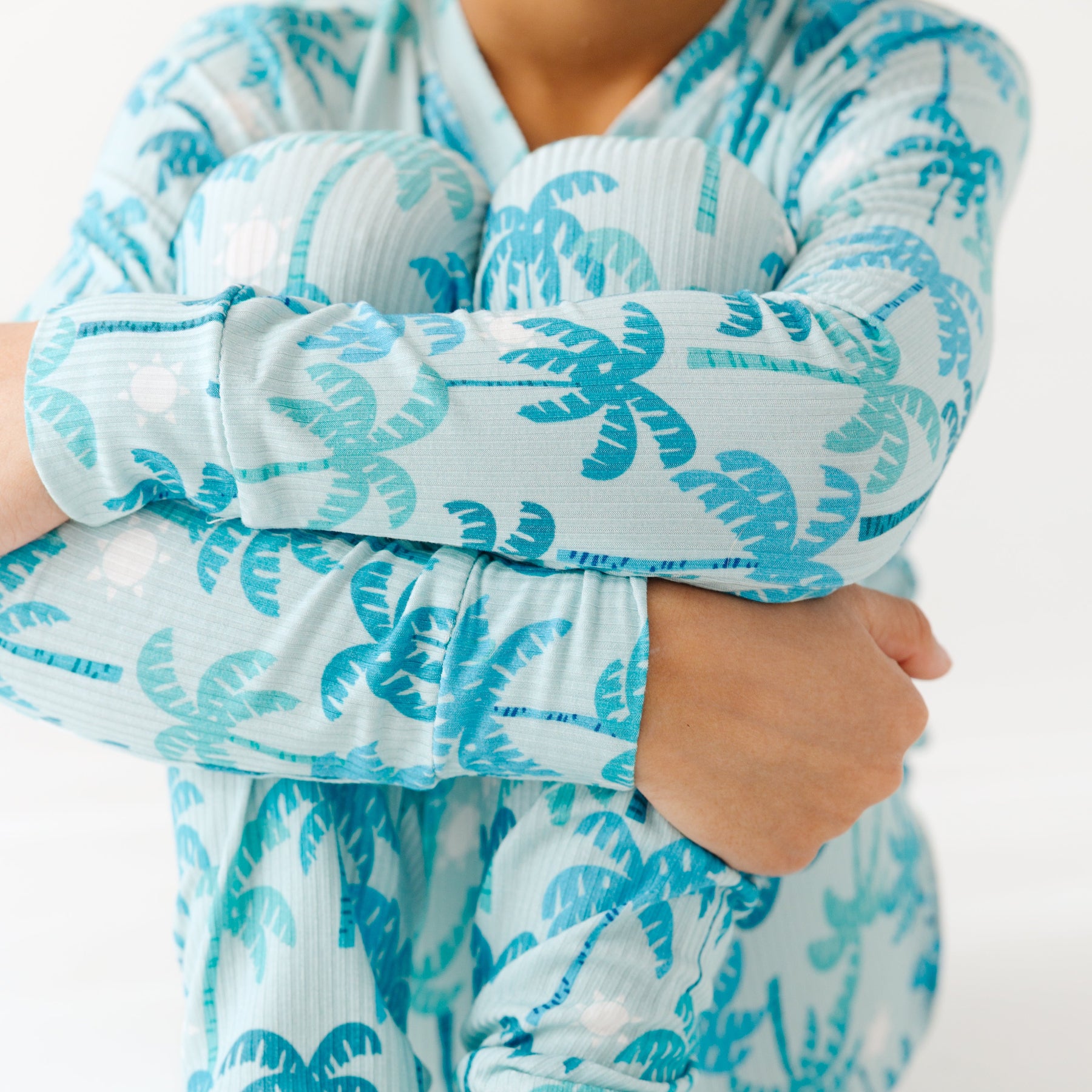 Close up of a child wearing a blue palm tree patterned pajama set on a white background.