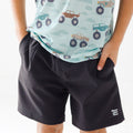Close up shot of a child wearing a light blue shirt with vehicle prints and black shorts on a white background