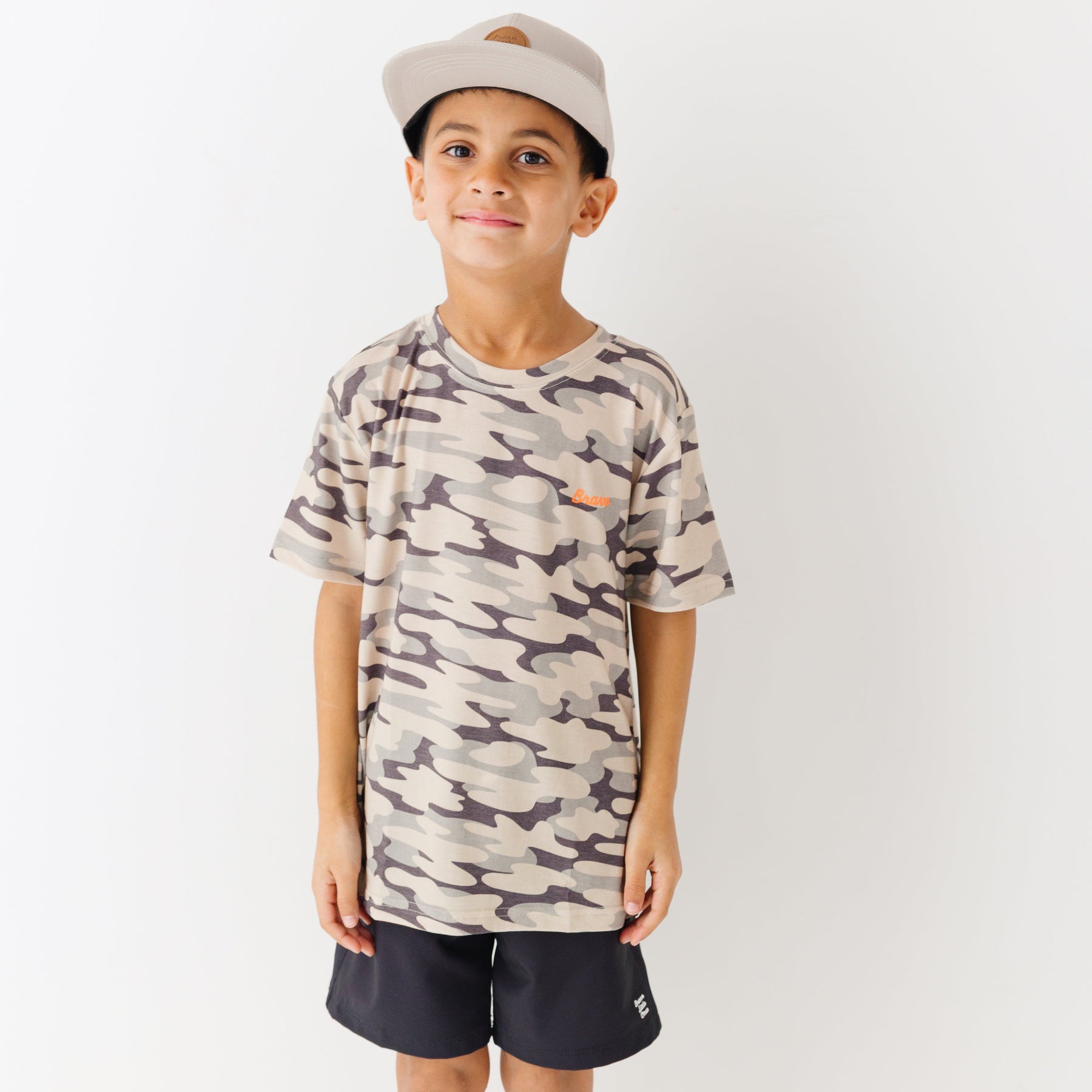 Child wearing a camouflage t-shirt, taupe cap and black shorts on a white background