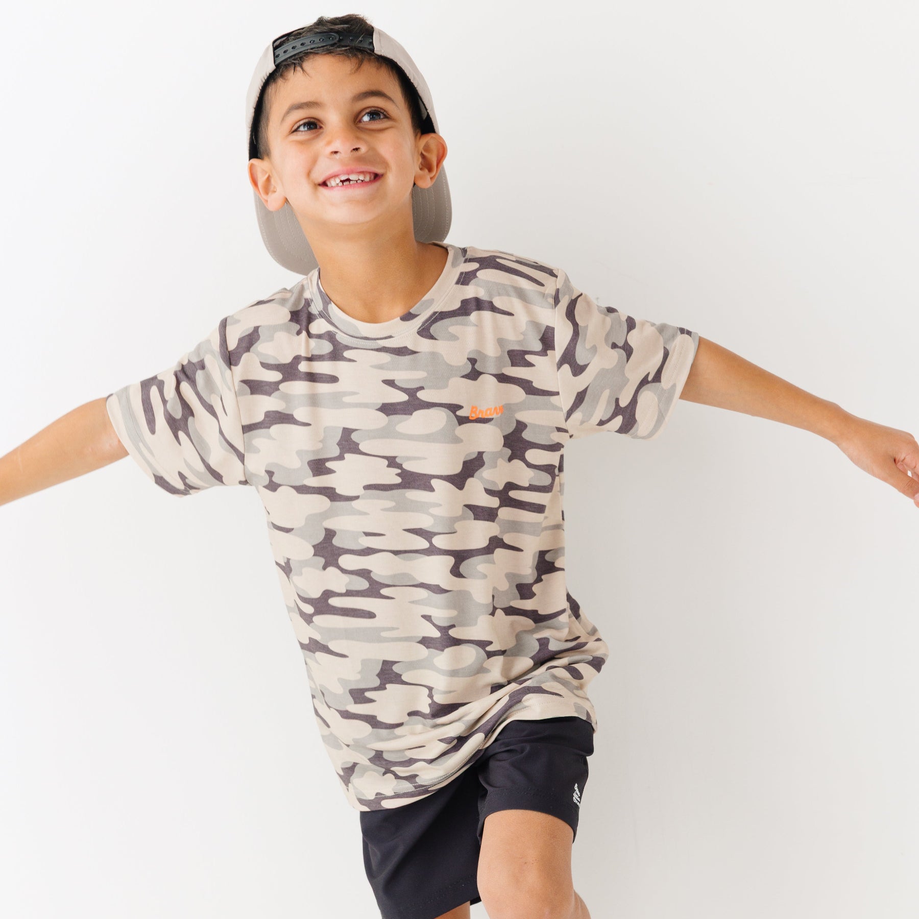 Child wearing a camouflage t-shirt, taupe cap and black shorts on a white background