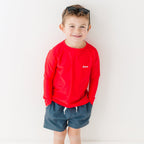 Child wearing a red rash guard and dark blue shorts on a white background