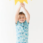 Child wearing a blue palm tree patterned outfit with arms raised holding a sun artwork against a white background