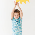 Child wearing a blue palm tree patterned outfit with arms raised holding a sun artwork against a white background