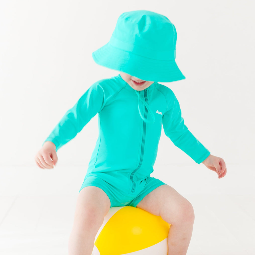 Child wearing a turquoise boy swimsuit and matching bucket hat with a yellow beach ball on a white background