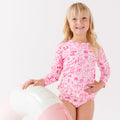 Child wearing a pink and white patterned long sleeve two-piece swimsuit on a white background holding a pink floatie